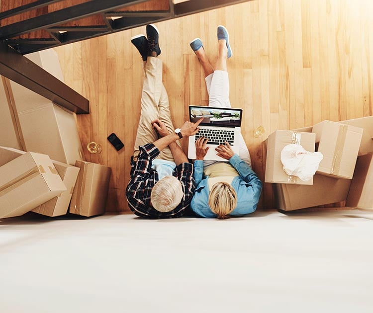 Couple sitting against a wall with boxes in new home viewing a laptop from above.