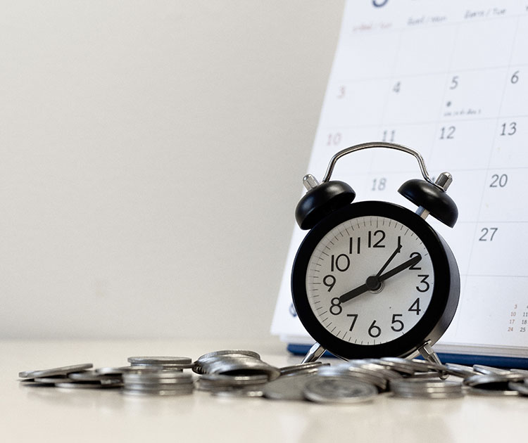 Photo of an alarm clock surrounded by coins with a calendar in the background.