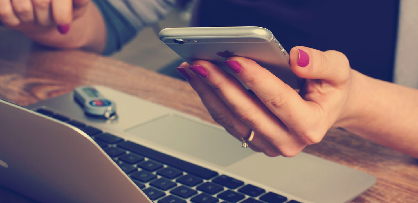 Hands use a mobile device with a desktop computer in the background.