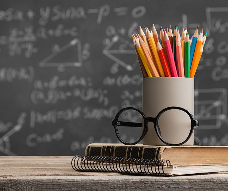 Books, pencils and glasses on desk with a chalkboard in the background.